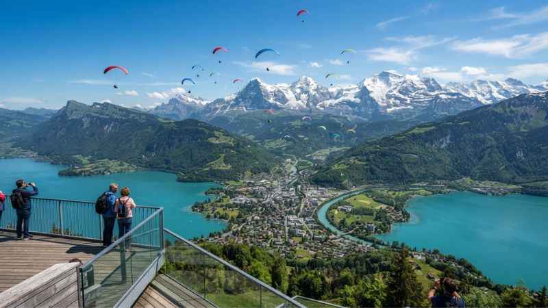 Panoramic view of Interlaken from Harder Kulm with paragliders flying above Lake Thun, Lake Brienz, and the Swiss Alps.